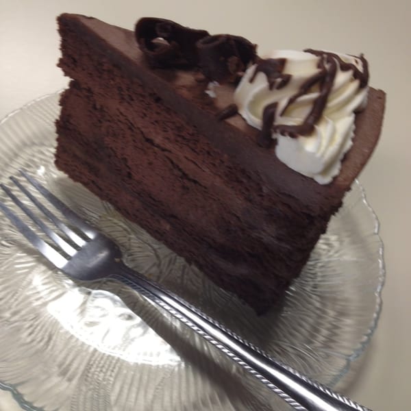A slice of rich chocolate cake with whipped cream and chocolate drizzle on a clear plate with a fork.